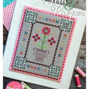 May include: A framed cross-stitch pattern with a floral design in shades of red, pink, and green. The pattern is titled "Prim & Proper" and is by Lori Holt of Bee in my Bonnet Co.