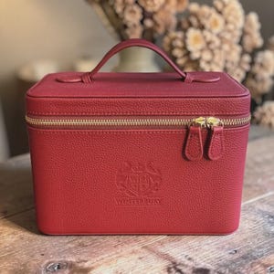 May include: A rectangular, deep red vanity case with a textured surface, a top handle, and a gold-toned zipper closure. The front features an embossed crest logo with the text WINTERBURY centered below it. The case has a structured, boxy shape.
