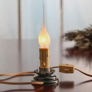 May include: A small electric candle lamp featuring a tan, drip-style plastic candle sleeve, a dark metal scalloped base, and a flame-shaped light bulb. The lamp includes a beige power cord with an inline thumb-wheel switch for operation.