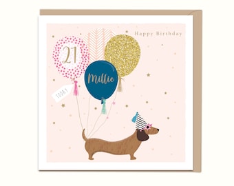 May include: A pink and gold birthday card with a dachshund wearing a party hat and sunglasses. The card says "Happy Birthday" and has balloons that say "21" and "Millie".