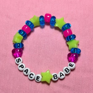 Op de afbeelding: Een kleurrijke kralenarmband met de tekst "SPACE BABE". De armband heeft neon groene ster- en hartvormige kralen, roze en blauwe kralen en witte letterkralen. De achtergrond is roze.
