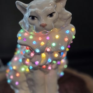 May include: A white ceramic cat figurine decorated with numerous small, glowing multi-colored LED lights. The cat has a glossy finish, pointed ears, and dark eyes, with the colorful lights wrapped around its body, creating a bright, illuminated effect.