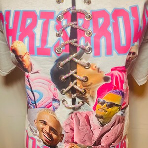 May include: White t-shirt with a pink and blue graphic print of Chris Brown and the text "Chris Brown" and "1-11 Tour". The shirt has a lace-up back with rhinestones.