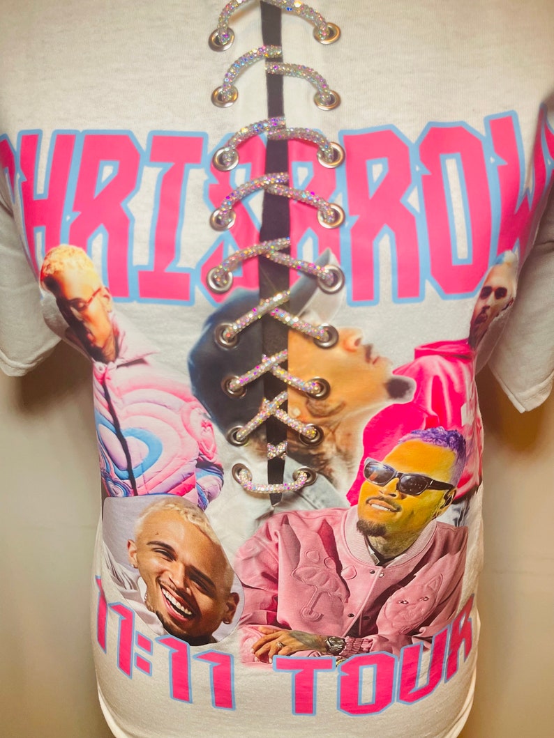 May include: White t-shirt with a pink and blue graphic print of Chris Brown and the text "Chris Brown" and "1-11 Tour". The shirt has a lace-up back with rhinestones.