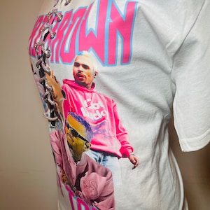 May include: White t-shirt with a pink and blue "Chris Brown Tour" graphic. The graphic features a photo of Chris Brown and another person. The shirt has a lace-up detail at the neckline.