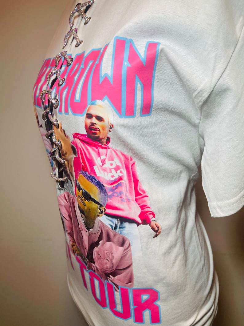 May include: White t-shirt with a pink and blue "Chris Brown Tour" graphic. The graphic features a photo of Chris Brown and another person. The shirt has a lace-up detail at the neckline.