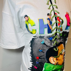 Pu&ograve; includere: Maglietta bianca con una stampa grafica verde, viola e gialla della parola "CHRI" e una foto di un uomo. La maglietta ha un dettaglio di pizzo verde sul davanti. La grafica include anche il testo "11:11 tour".