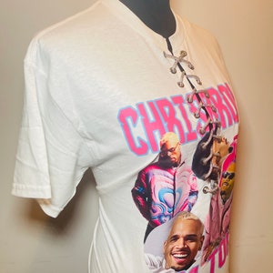 May include: White t-shirt with a lace-up detail at the neckline. The shirt features a graphic print of Chris Brown with the text "Chris Brown 11:11 Tour".