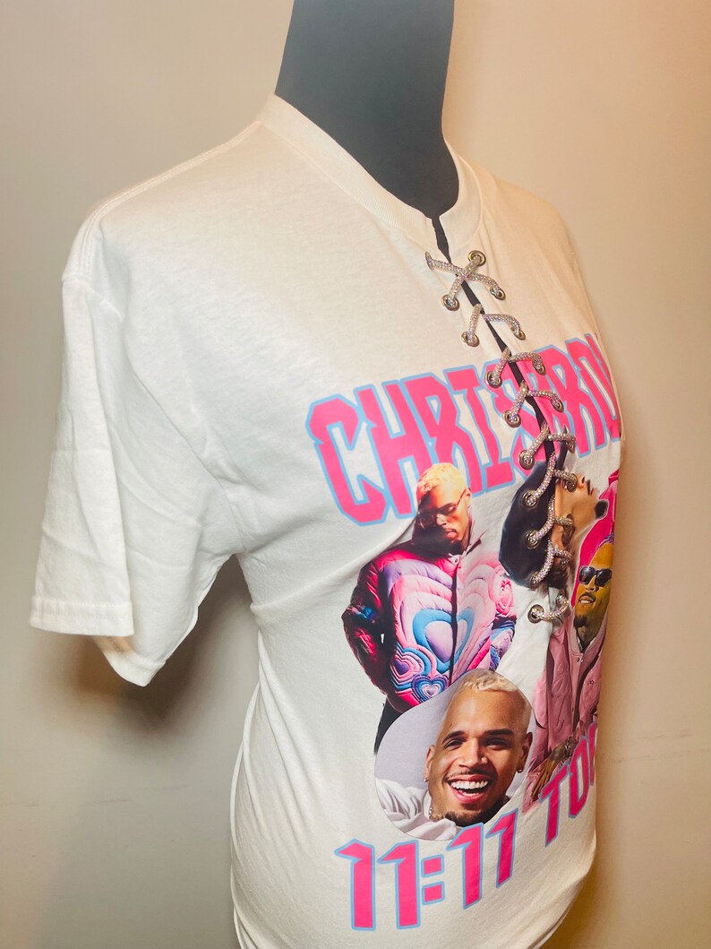 May include: White t-shirt with a lace-up detail at the neckline. The shirt features a graphic print of Chris Brown with the text "Chris Brown 11:11 Tour".
