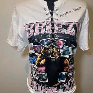 May include: White t-shirt with a graphic of a person holding a microphone, surrounded by photo-like images. The word "BREEZY" is in pink letters with a black outline. The shirt has a lace-up detail at the neckline. The text "BOWL XX" is at the bottom.