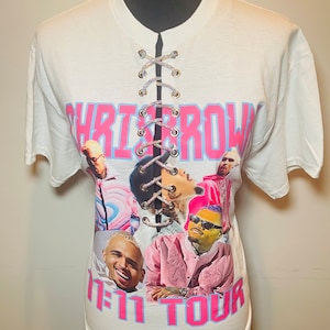May include: White t-shirt with a pink and blue graphic print of Chris Brown and the text "1111 Tour". The shirt has a lace-up detail in the front.