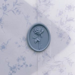 May include: An oval-shaped light blue wax seal featuring a raised relief design of a rose flower attached to the top of a skeleton key. The seal is centered on a translucent white envelope decorated with a faint blue floral pattern.