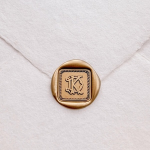 May include: A gold wax seal with an intricate monogram, positioned on a white envelope. The seal is square with a decorative edge, and the monogram features two intertwined letters. The envelope is made of textured paper.