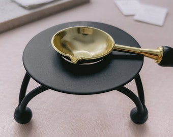 May include: A black metal wax warmer with a gold-colored spoon-shaped wax melting ladle. The ladle has a wooden handle. The warmer has a round top and three curved legs with ball feet. The background is a light pink surface.