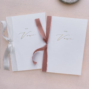A set of 2 white vow books gold foil printed with His Vows and Her Vows in calligraphy script, tied in rose colored and white silk ribbon