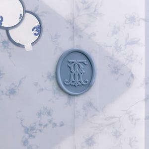 May include: A light blue wax seal featuring an embossed monogram of the letters TR, centered on a translucent vellum envelope with a delicate blue floral pattern. Two circular adhesive backing stickers are visible in the upper left corner.