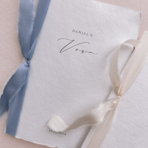 Set of 2 white handmade paper vow books printed with personalized names and date, tied in dusty blue and off-white silk ribbons