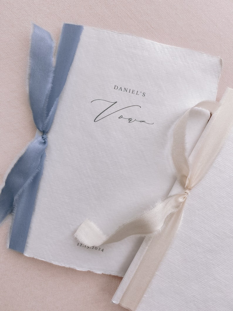 Set of 2 white handmade paper vow books printed with personalized names and date, tied in dusty blue and off-white silk ribbons