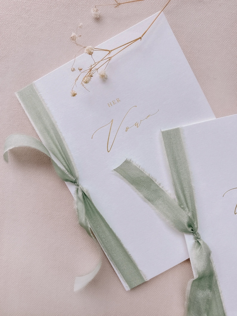 A set of 2 white vow books gold foil printed with His Vows and Her Vows in calligraphy script, tied in sage green silk ribbon