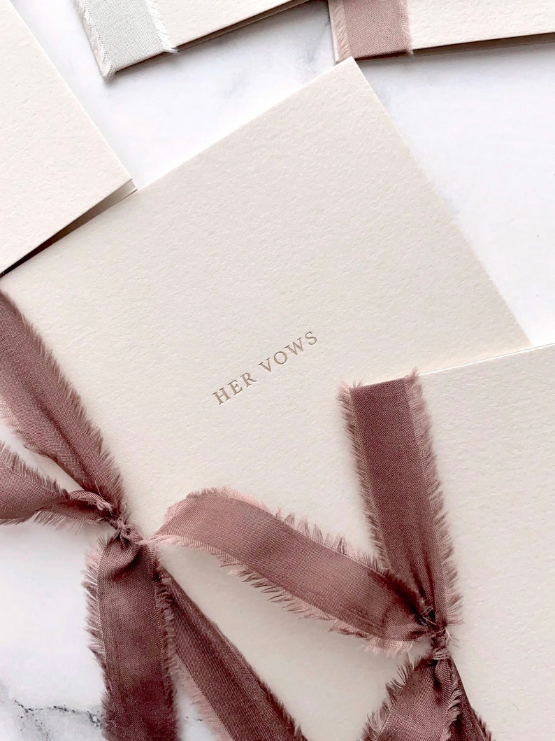 A set of 2 minimalist beige vow books gold foil printed with His Vows and Her Vows, tied in burgundy colored silk ribbons