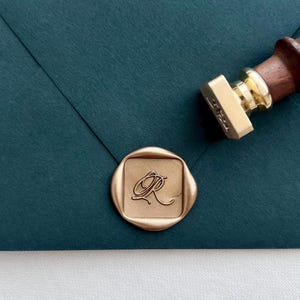 Square-shaped custom design gold wax seal featuring an initial in elegant calligraphy script, on a dark green envelope