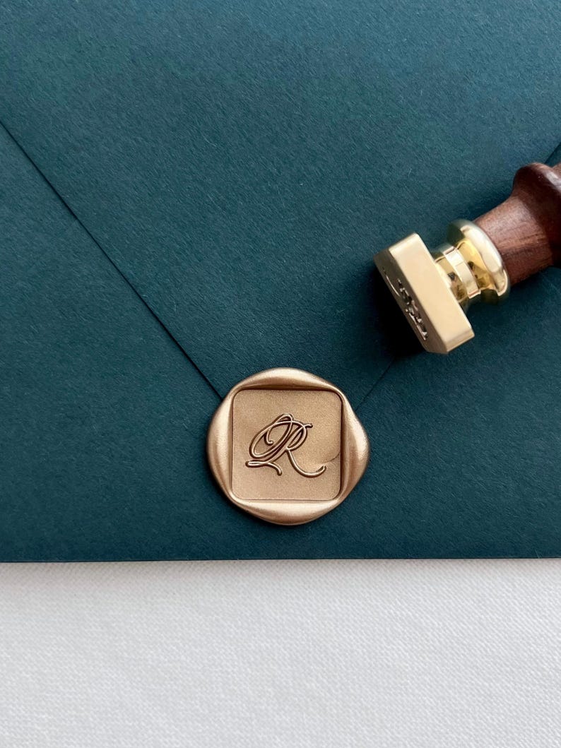 Square-shaped custom design gold wax seal featuring an initial in elegant calligraphy script, on a dark green envelope