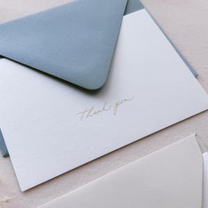 gold embossed thank you card featuring thank you in modern calligraphy script with a dusty blue envelope