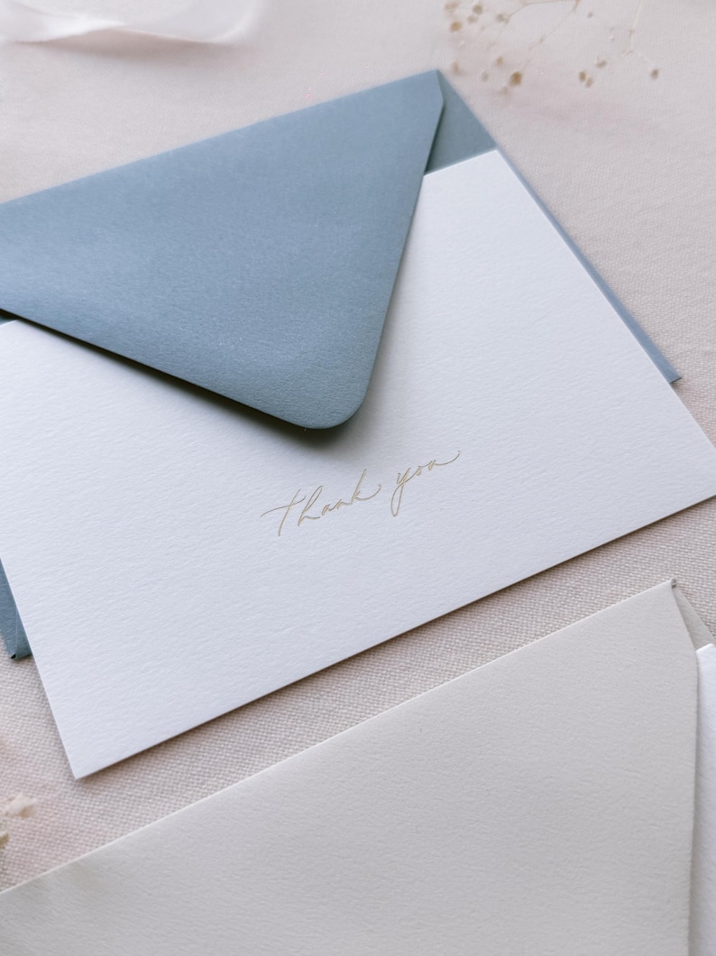 gold embossed thank you card featuring thank you in modern calligraphy script with a dusty blue envelope