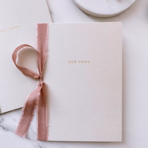 A set of beige vow books with His Vows and Her Vows gold foil printed on the cover, tied in rose colored silk ribbon