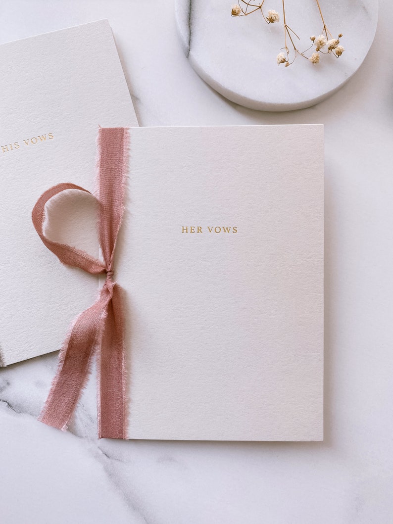 A set of beige vow books with His Vows and Her Vows gold foil printed on the cover, tied in rose colored silk ribbon