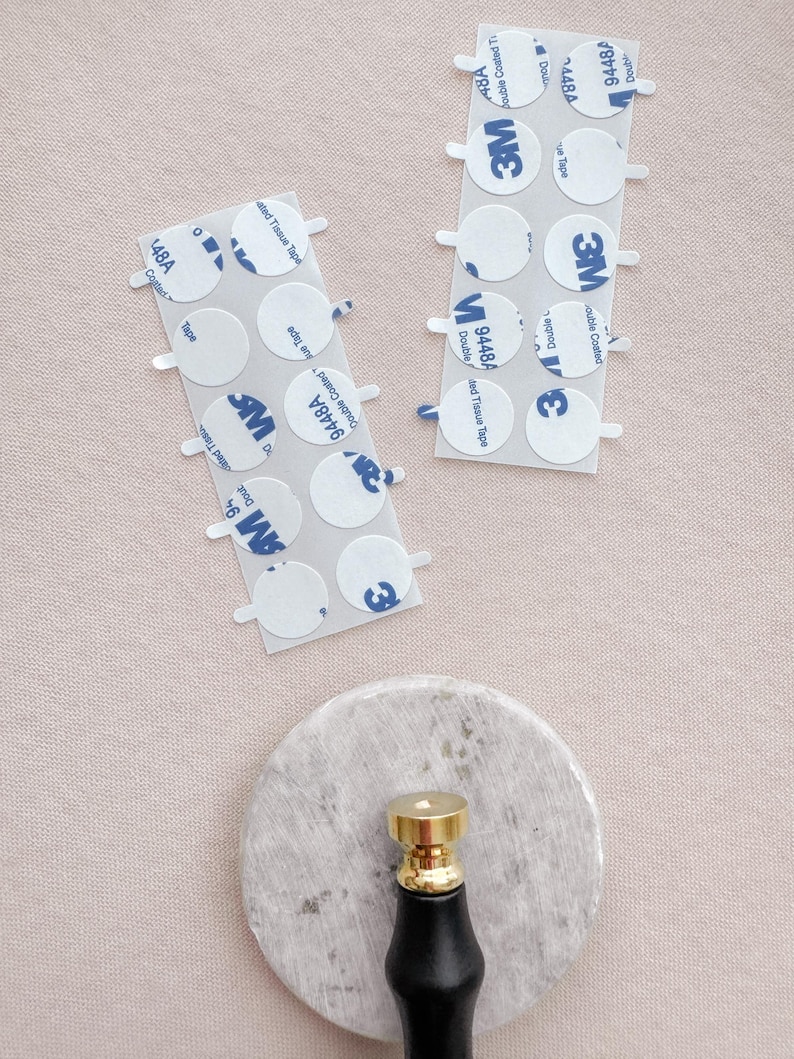 Pu&ograve; includere: Due fogli di nastro adesivo a doppio lato 3M con pi&ugrave; cerchi adesivi rotondi. Il nastro &egrave; etichettato con il numero di prodotto 9448A e 4876. Il nastro &egrave; su una superficie rosa. Un timbro di sigillo di cera dorato e nero &egrave; su una superficie di marmo bianco.