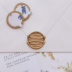 Könnte beinhalten: Ein goldenes Wachssiegel mit welliger, gerippter Textur, zentriert auf einem weißen Umschlag. Zwei weitere goldene Wachssiegel mit weißer 3M-Klebefläche befinden sich darüber. Eine Einladungskarte mit dem Text 23.II.202 at half past five, The Langham Estate, Napa, California, reception to follow ist zu sehen.