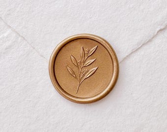 May include: A gold-colored wax seal with a botanical leaf design. The seal is circular and embossed with a detailed illustration of a sprig with leaves and small buds. The seal is set against a white paper background.