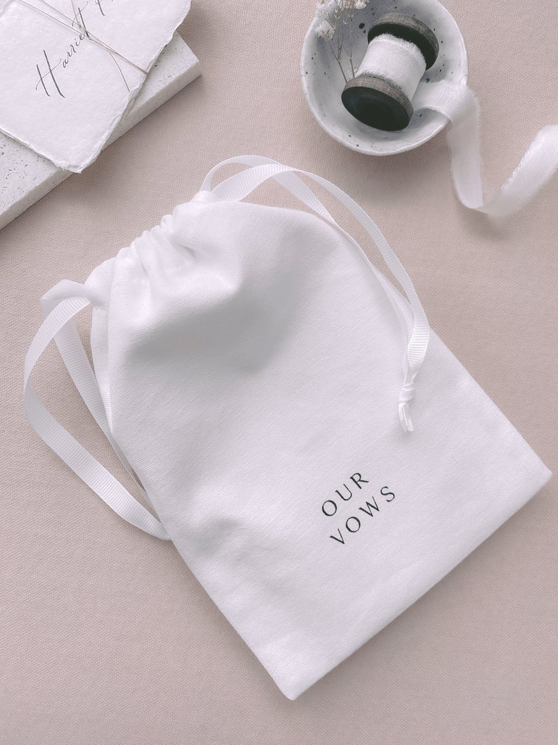 White keepsake pouch in white drawstring with OUR VOWS printed on it in black ink