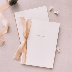 A set of 2 beige vow books gold foil printed with His Vows and Her Vows, tied in gold colored silk ribbons
