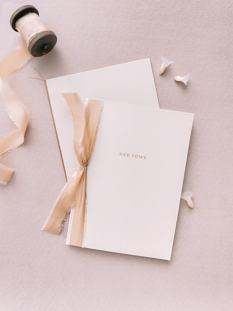 A set of 2 beige vow books gold foil printed with His Vows and Her Vows, tied in gold colored silk ribbons