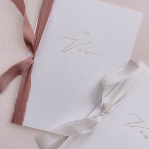 A set of 2 white vow books gold foil printed with His Vows and Her Vows in calligraphy script, tied in rose colored and white silk ribbon