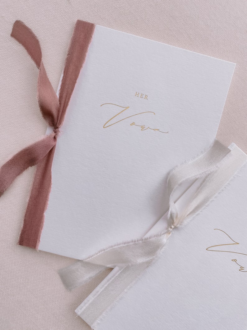 A set of 2 white vow books gold foil printed with His Vows and Her Vows in calligraphy script, tied in rose colored and white silk ribbon