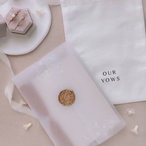 Vow books packed inside white floral vellum jacket, adorned with a gold wax seal, placed next to a white keepsake linen pouch
