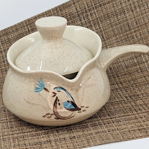 Puede incluir: Una cacerola de cerámica beige con tapa y asa. La cacerola está decorada con un diseño de pájaro azul y marrón. La tapa y el cuerpo de la cacerola tienen un patrón moteado.