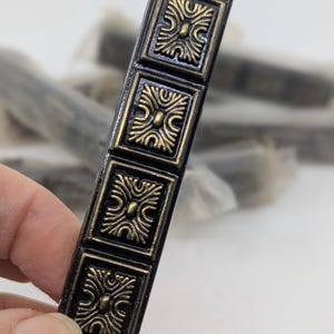 May include: A black and gold cabinet pull with a rectangular shape. The pull features five square panels with an ornate, symmetrical design. The gold detailing contrasts with the black background. Other pulls are visible in the background.