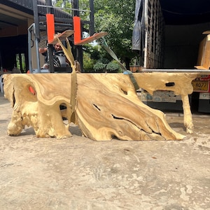 Puede incluir: Dos piezas grandes de madera natural, probablemente una tapa de mesa, con una forma orgánica única y un color marrón claro. La madera tiene muchos nudos y patrones de veta. La madera se está cargando en un camión.