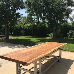 Puede incluir: Una gran encimera de mesa de madera natural con detalles de borde vivo, apoyada en un bastidor rodante de metal. La encimera de la mesa es de un color marrón intenso con un acabado liso y pulido. La mesa está al aire libre sobre una superficie de grava.