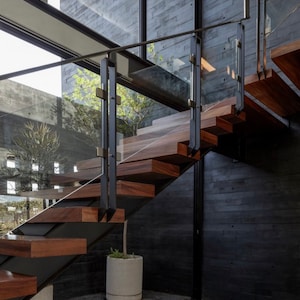 Może przedstawiać: Nowoczesne schody z drewnianymi stopniami i szklaną balustradą. Schody znajdują się w domu z betonową ścianą i dużymi oknami.