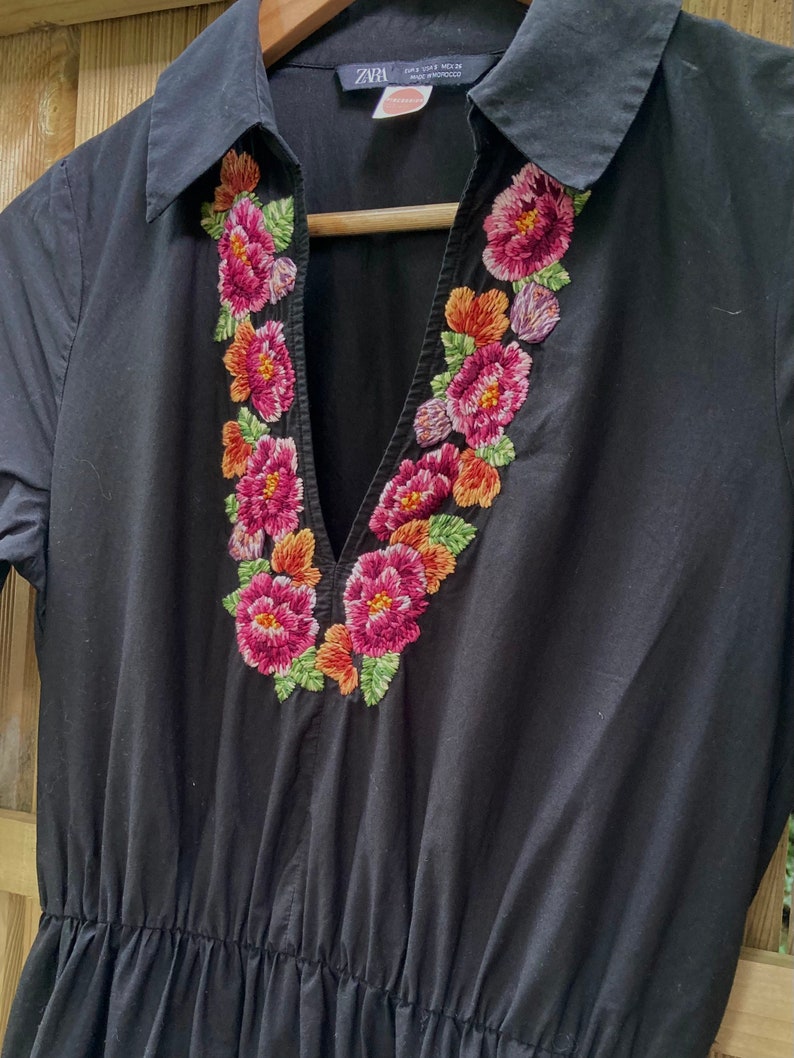 May include: Black short-sleeved dress with a V-neck and embroidered floral detail around the neckline. The dress has a gathered waist and is made by Zara.