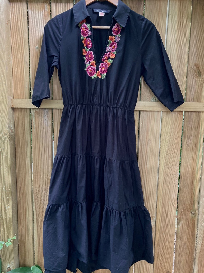 May include: A black tiered midi dress with a collared neckline and embroidered floral detail around the neckline. The dress has a v-neck and 3/4 sleeves.
