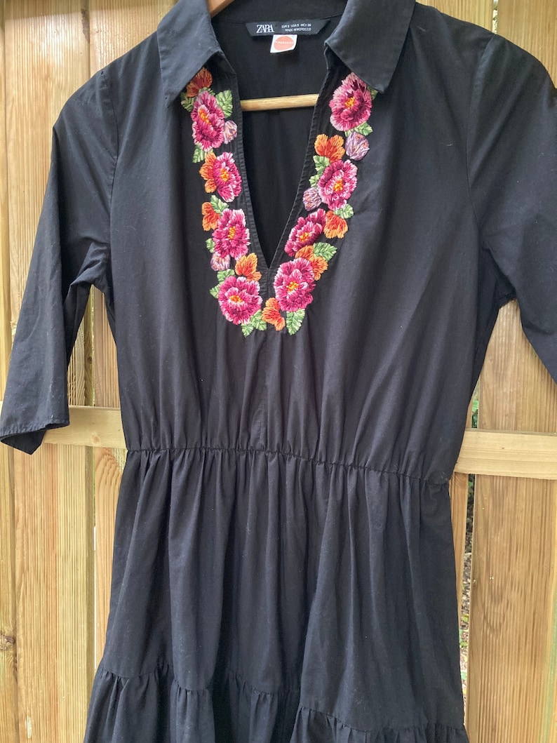 May include: A black short-sleeved dress with a V-neck and a floral embroidered neckline. The dress has a tiered skirt and a collared neckline. The embroidery is in shades of pink, orange, and green.