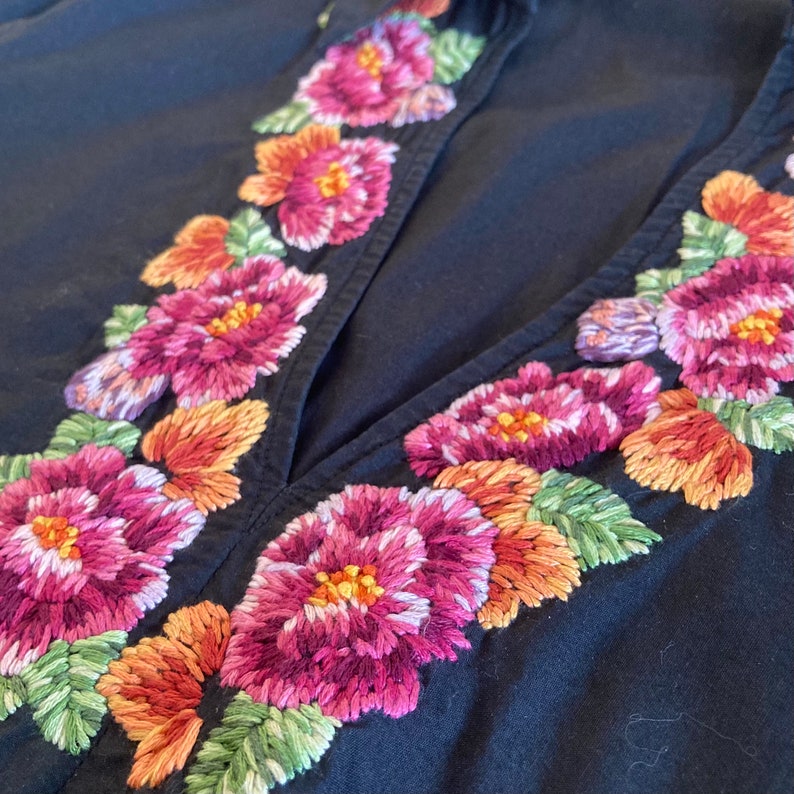 May include: A black cotton shirt with embroidered flowers in shades of pink, orange, and green around the neckline.