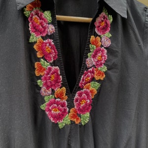 May include: Black collared shirt with a V-neckline and embroidered floral design around the neckline. The embroidery features pink, red, orange, and green flowers.