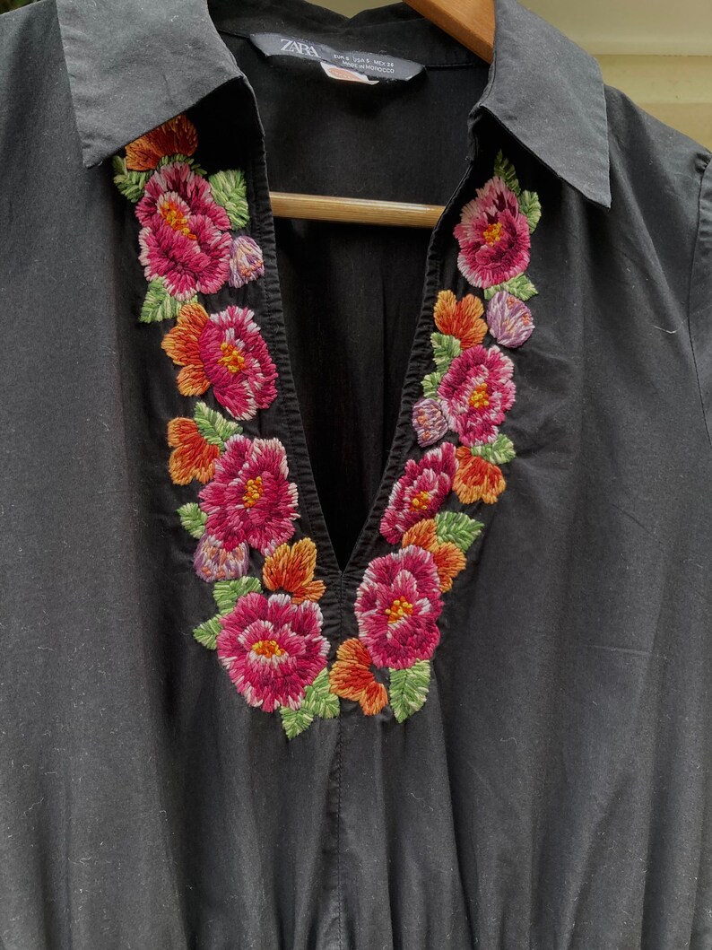 May include: Black collared shirt with a V-neckline and embroidered floral design around the neckline. The embroidery features pink, red, orange, and green flowers.